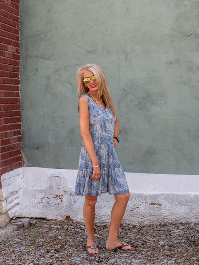 A model wearing a blue sleeveless mesh tiered shift dress that has faux buttons and a floral pattern. She has it on with brown sandals.