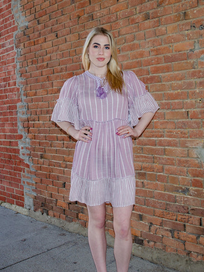 Model is wearing a striped purple and white half sleeve dress that is thigh length and cinches at waist. The neckline has string details to allows for adjusting of the open neck. 
