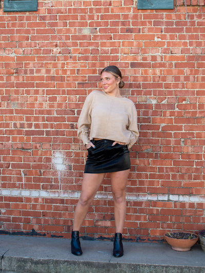 A model wearing a black faux leather mini skirt. The model has it paired with a tan sweater and black booties.