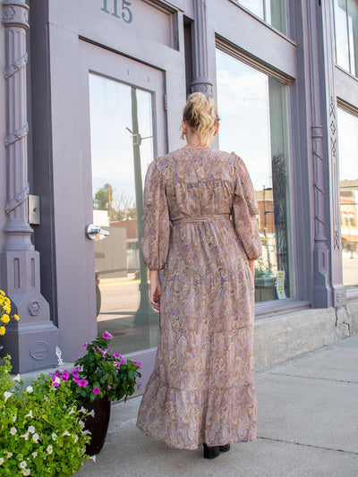 A model wearing a wrap style maxi dress with a light brown colored paisley print all over it. The model has it paired with black booties.