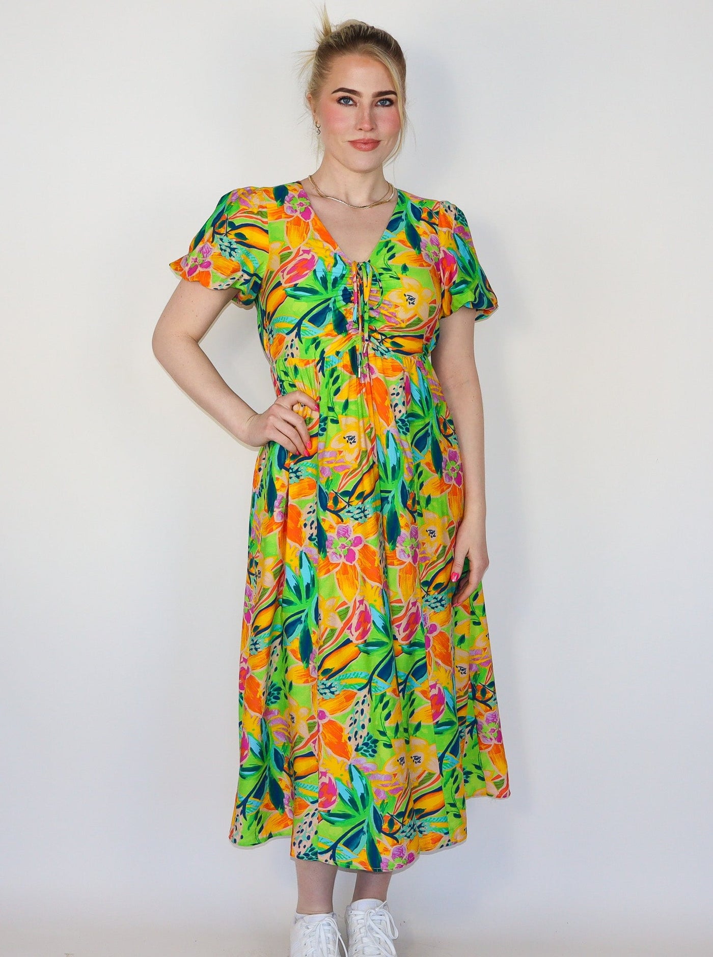 Model is wearing a flowy multi colored midi dress with puffy sleeves, a vneck, and string detail on the neckline. Dress is paired with white tennis shoes. 