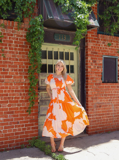 Model is wearing a orange and white floral printed maxi dress with puffed sleeves, cinching at boddess, and a tie to tie the waist. Worn with beige sandals.