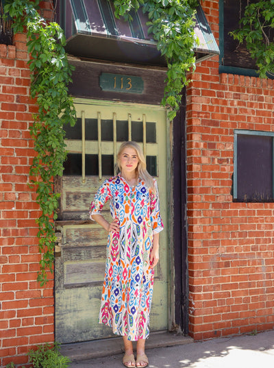 Summery abstract printed button up maxi dress with cuffed sleeves, collared neckline, and slights on each side. Worn with beige sandals.