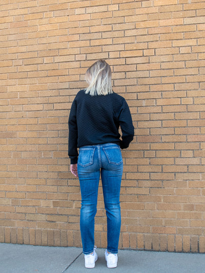 A model wearing a medium wash ankle skinny jean. The model has it paired with a black sweatshirt and white sneakers.