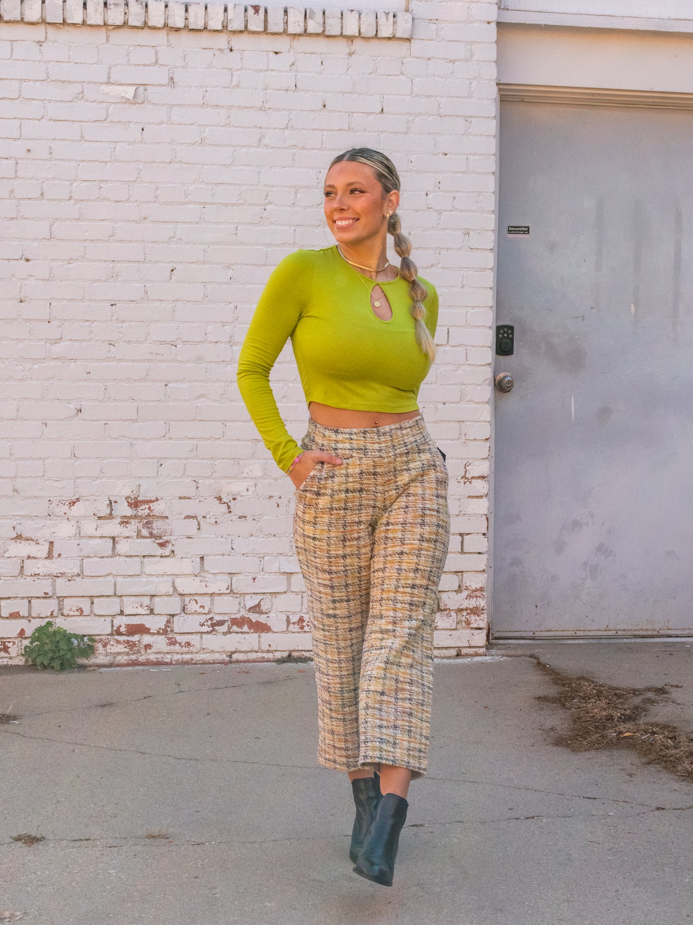 A model wearing a green long sleeve crop top with a cutout. She has it on with plaid boucle pants and black booties.