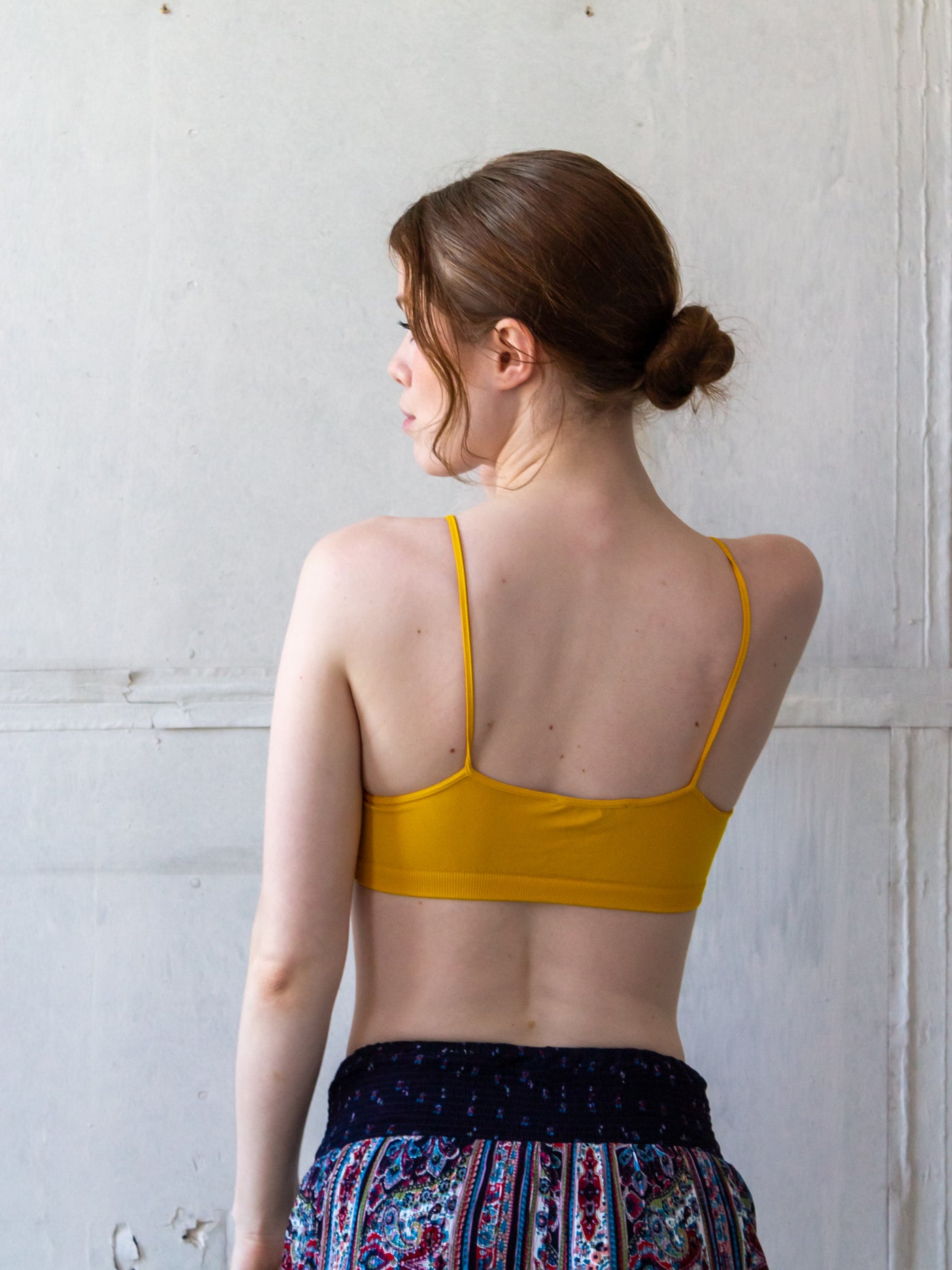 A model wearing a yellow bralette that is simple, stretchy material. It has two straps and a cage like design with two straps. Each strap is attached at the side seam and crosses over in front to attach to the other side about one inch from the small v neck. The main straps attach at the middle of each bra side and pull it up to form a peak.
