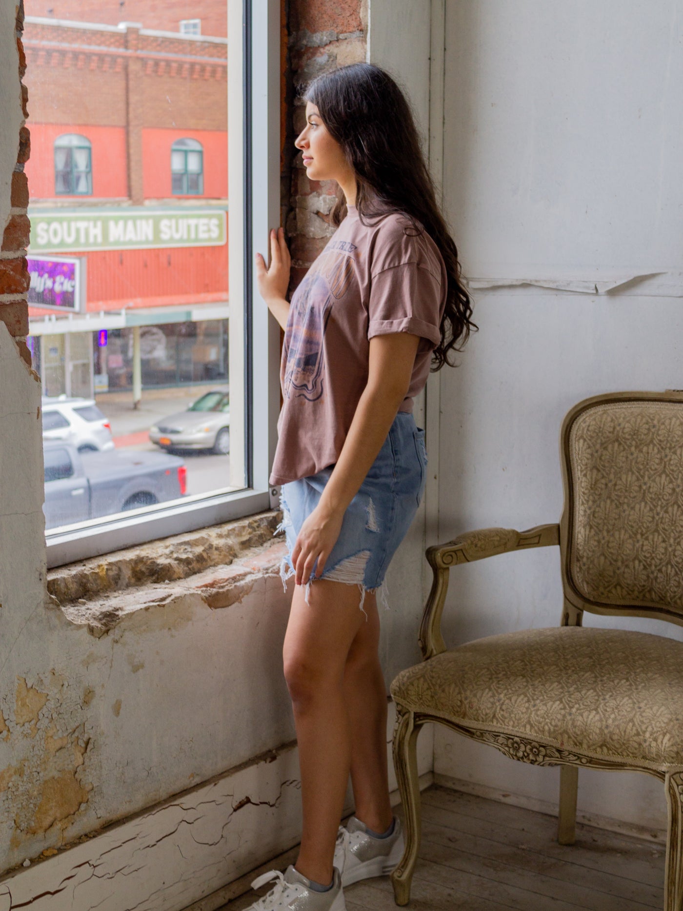 A model wearing a light wash mini skirt with a frayed hem and stressing throughout. The mini skirt is a mid rise. The model has it on with a graphic tee and white sneakers.