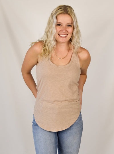 A model wearing a tan slub, relaxed fit tank top with a curved hem. Th model has it on with light wash denim jeans.