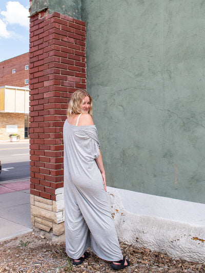 A model wearing a gray wide leg, oversized jumpsuit that has buttons to the waist. The model has paired it with a tan brami underneath and black sandals.