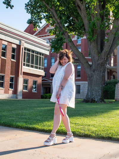 A model wearing a white sequin button down dress with white platform sandals and a pink wristlet purse. 