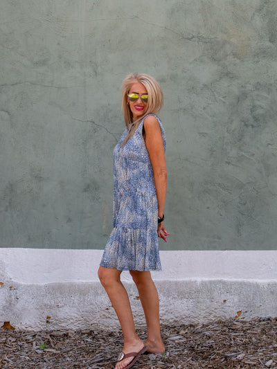 A model wearing a blue sleeveless mesh tiered shift dress that has faux buttons and a floral pattern. She has it on with brown sandals.