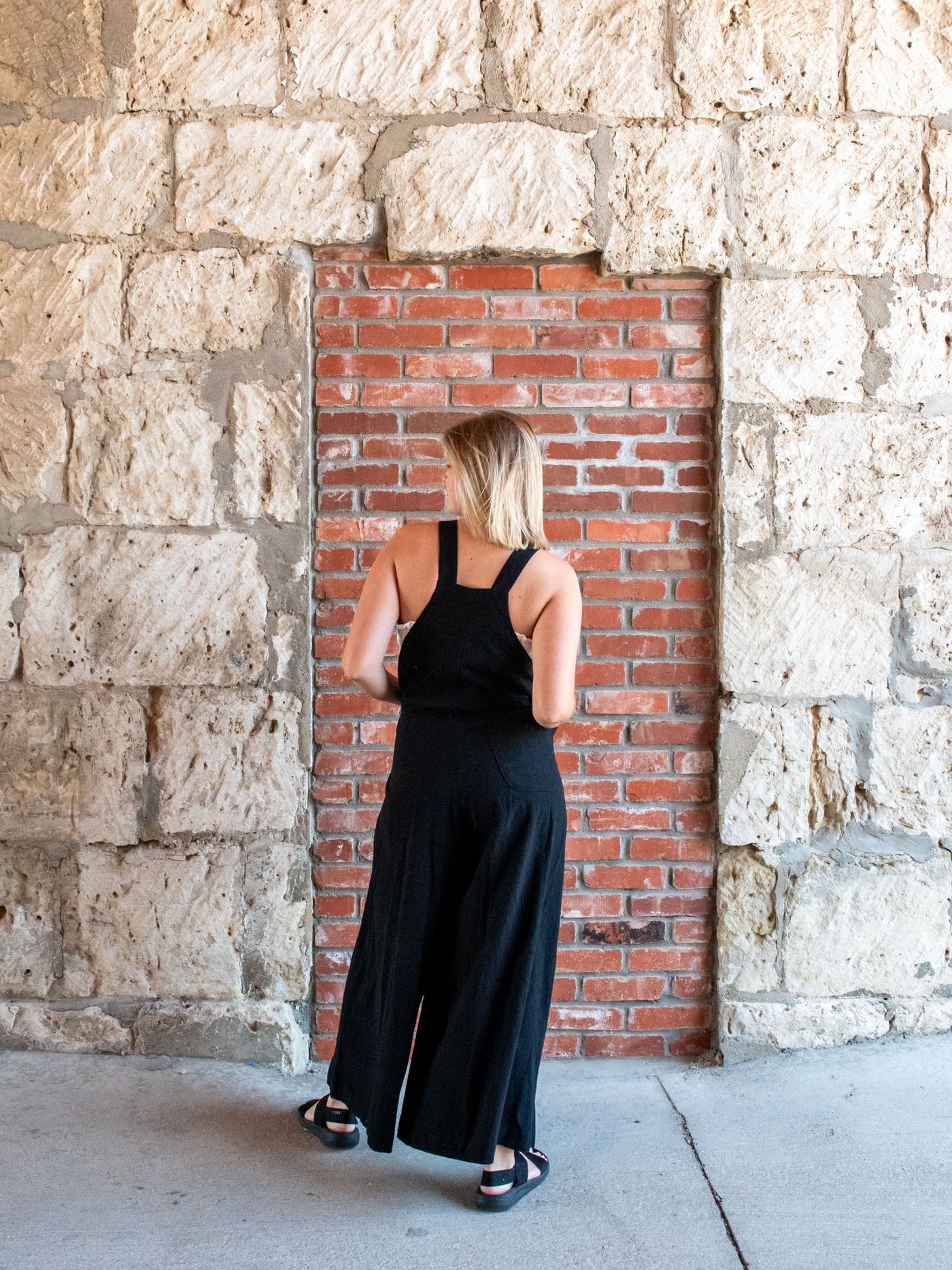 A model wearing a black linen jumpsuit that has three buttons and wide legs. She has it layered with a white tube top and black sandals.