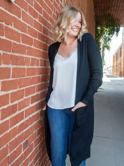 A model wearing a long, black open face cardigan. The model has it paired over a white cami and medium wash jeans.