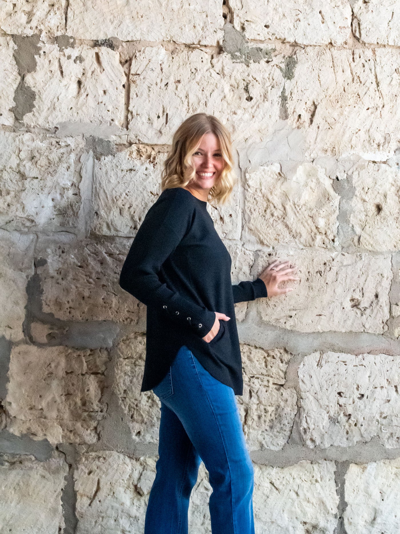 A model wearing a black sweater with a lace-up sleeve detail. The model has it paired with a medium wash straight leg jean.