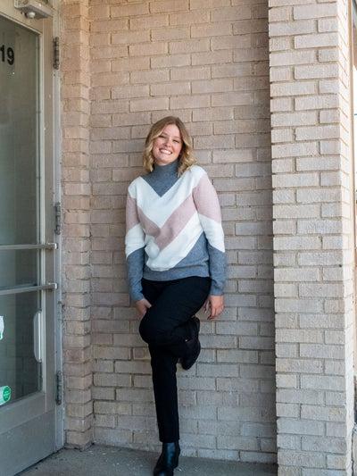 A model wearing a mock neck sweater with a gray, white, and blush chevron design. The model paired it with a pair of black pants and black boots.