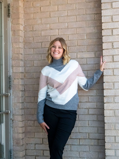A model wearing a mock neck sweater with a gray, white, and blush chevron design. The model paired it with a pair of black pants.