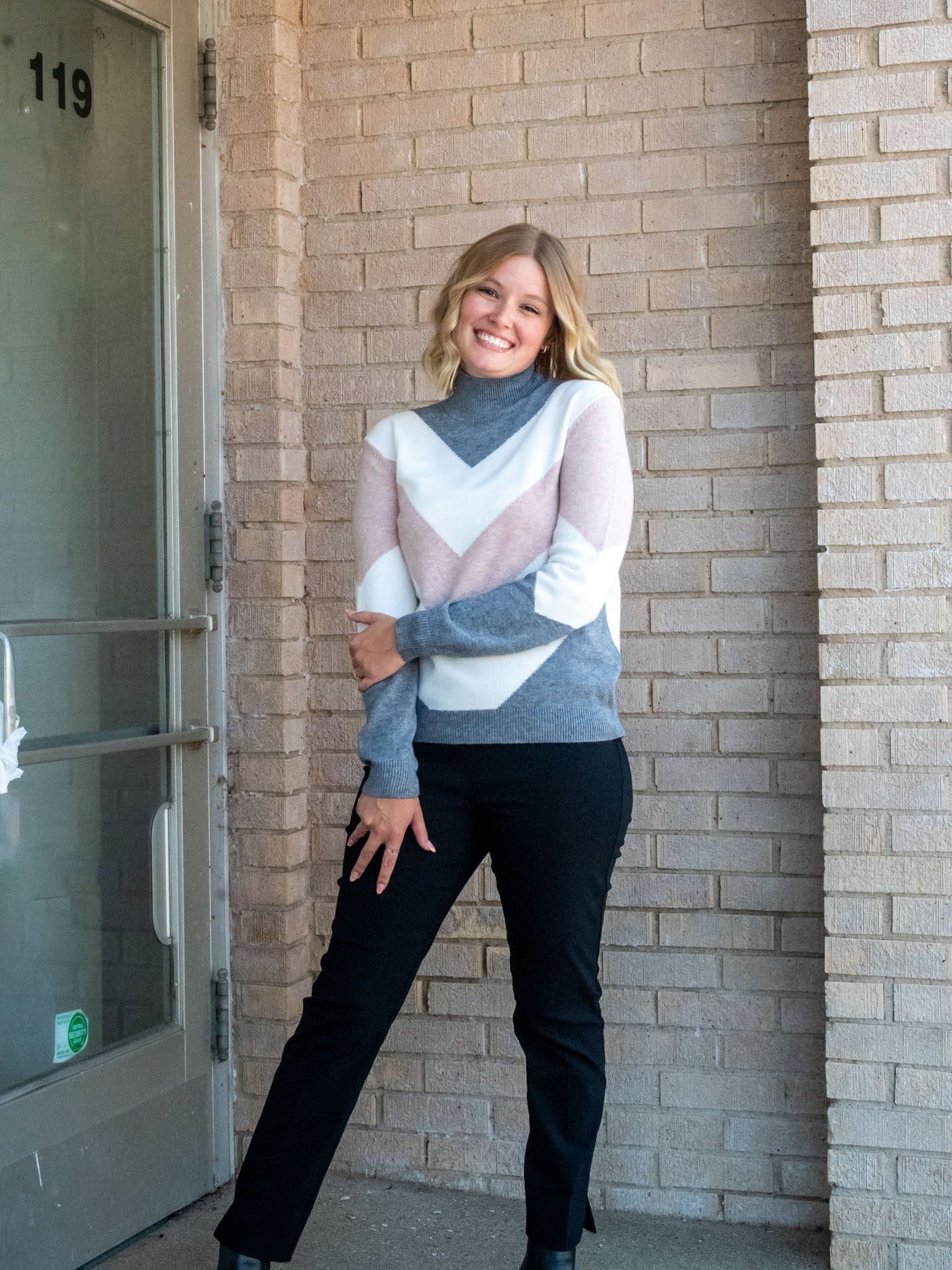 A model wearing a mock neck sweater with a gray, white, and blush chevron design. The model paired it with a pair of black pants.