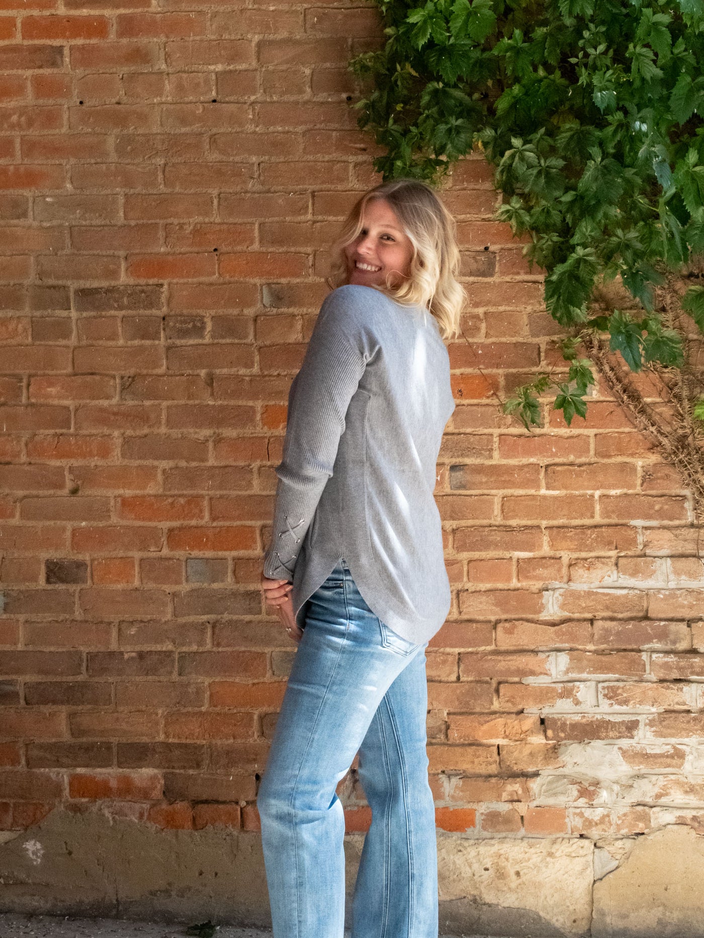 A model wearing a gray sweater with a lace-up sleeve detail. The model has it paired with a light wash straight leg jean.