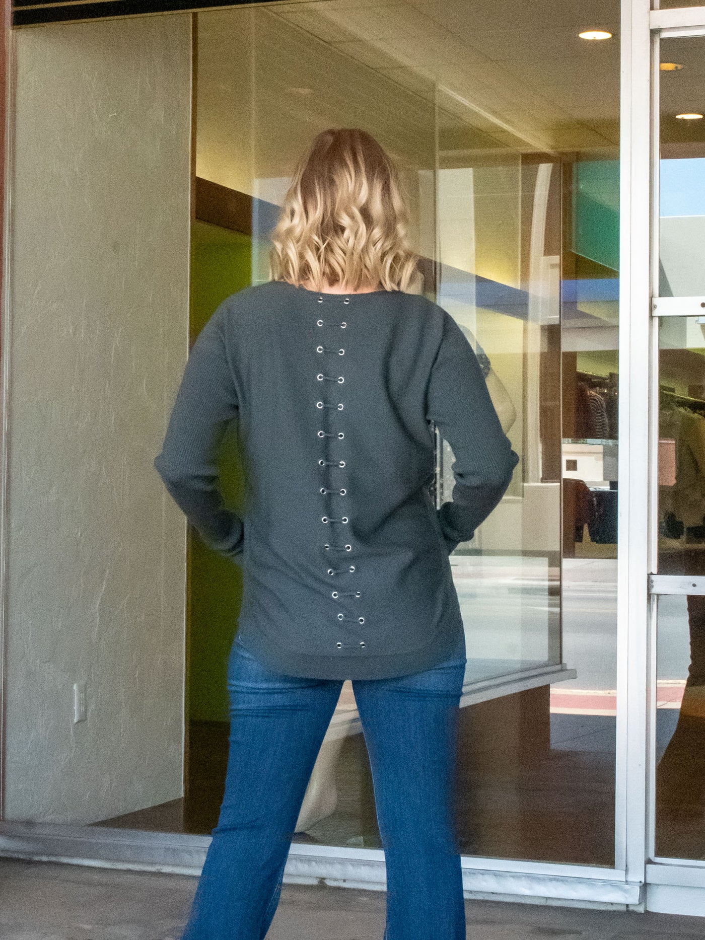 A model wearing an olive green knit sweater with a lace-up back detail. The model has it paired with a pair of dark wash, flared jeans.