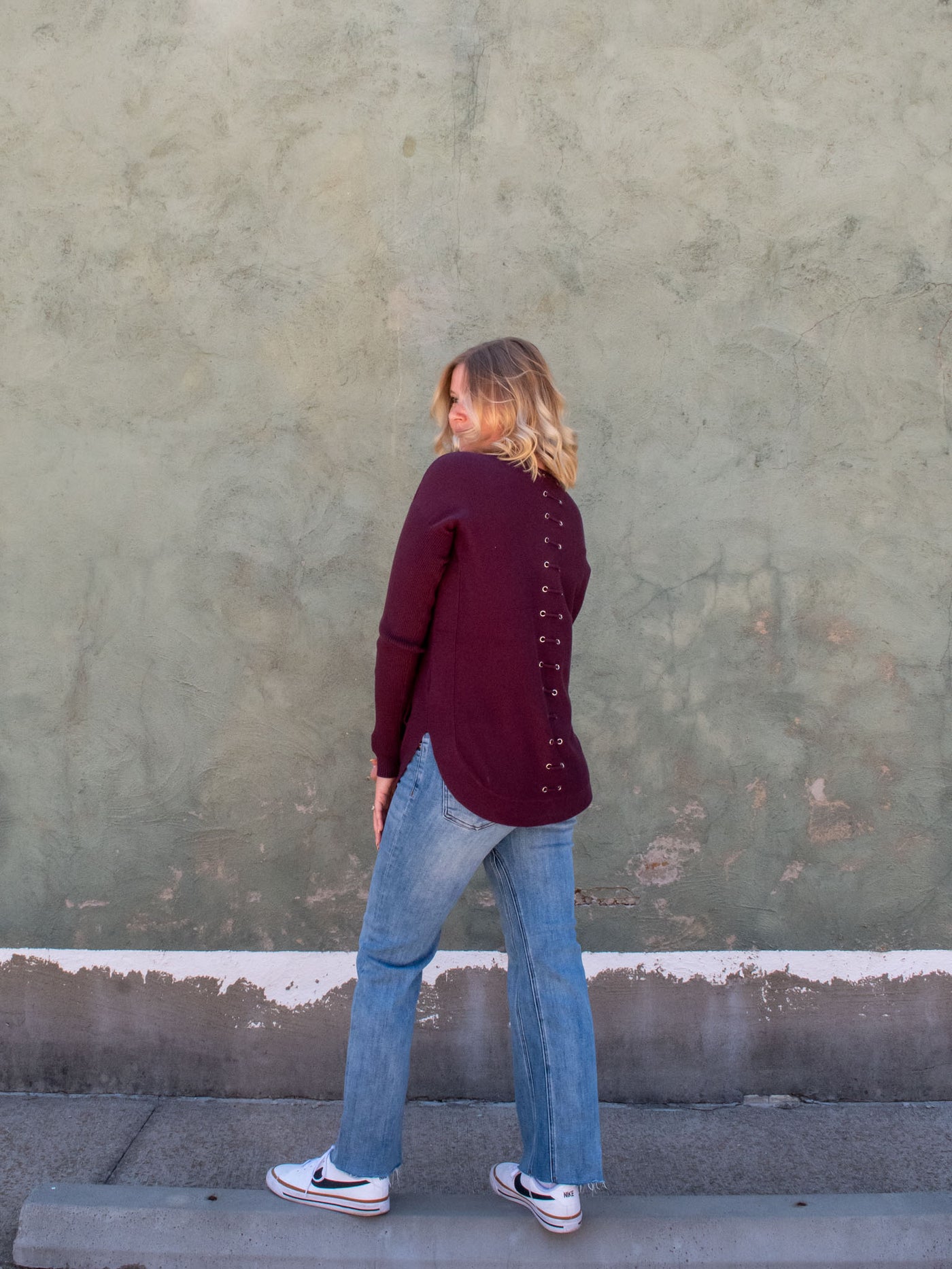 A model wearing a deep red knit sweater with a lace-up back detail. The model has it paired with a pair of light wash, straight jeans and white sneakers.