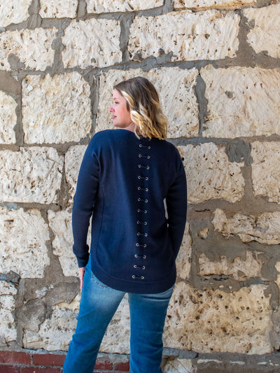 A model wearing a navy blue knit sweater with a lace-up back detail. The model has it paired with a pair of light wash, straight jeans.
