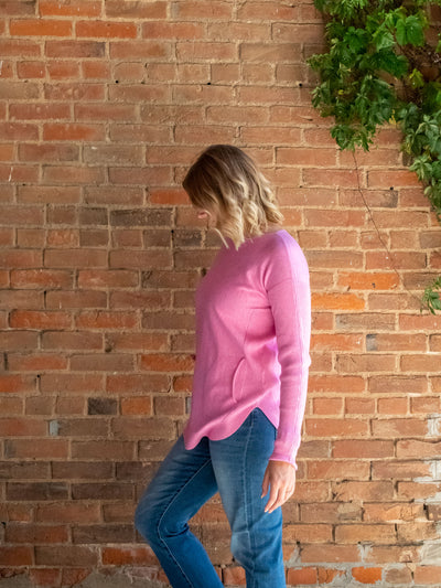 A model wearing a pink knit sweater with a lace-up back detail. The model has it paired with a pair of light wash, straight jeans.