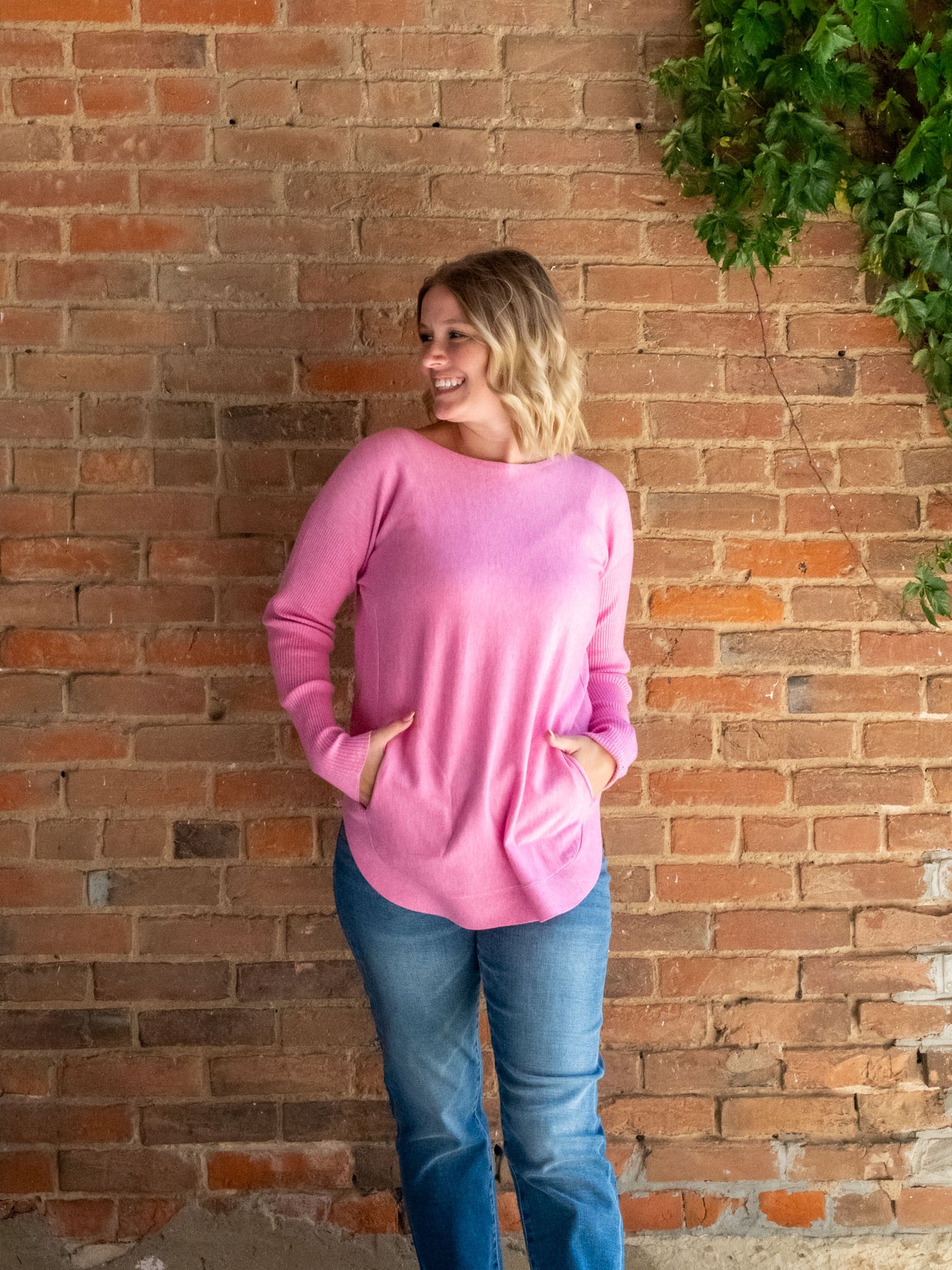 A model wearing a pink knit sweater with a lace-up back detail. The model has it paired with a pair of light wash, straight jeans.