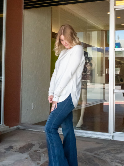 A model wearing a white sweater with a lace-up detail sleeve. The model has it paired with a dark wash flared jean.