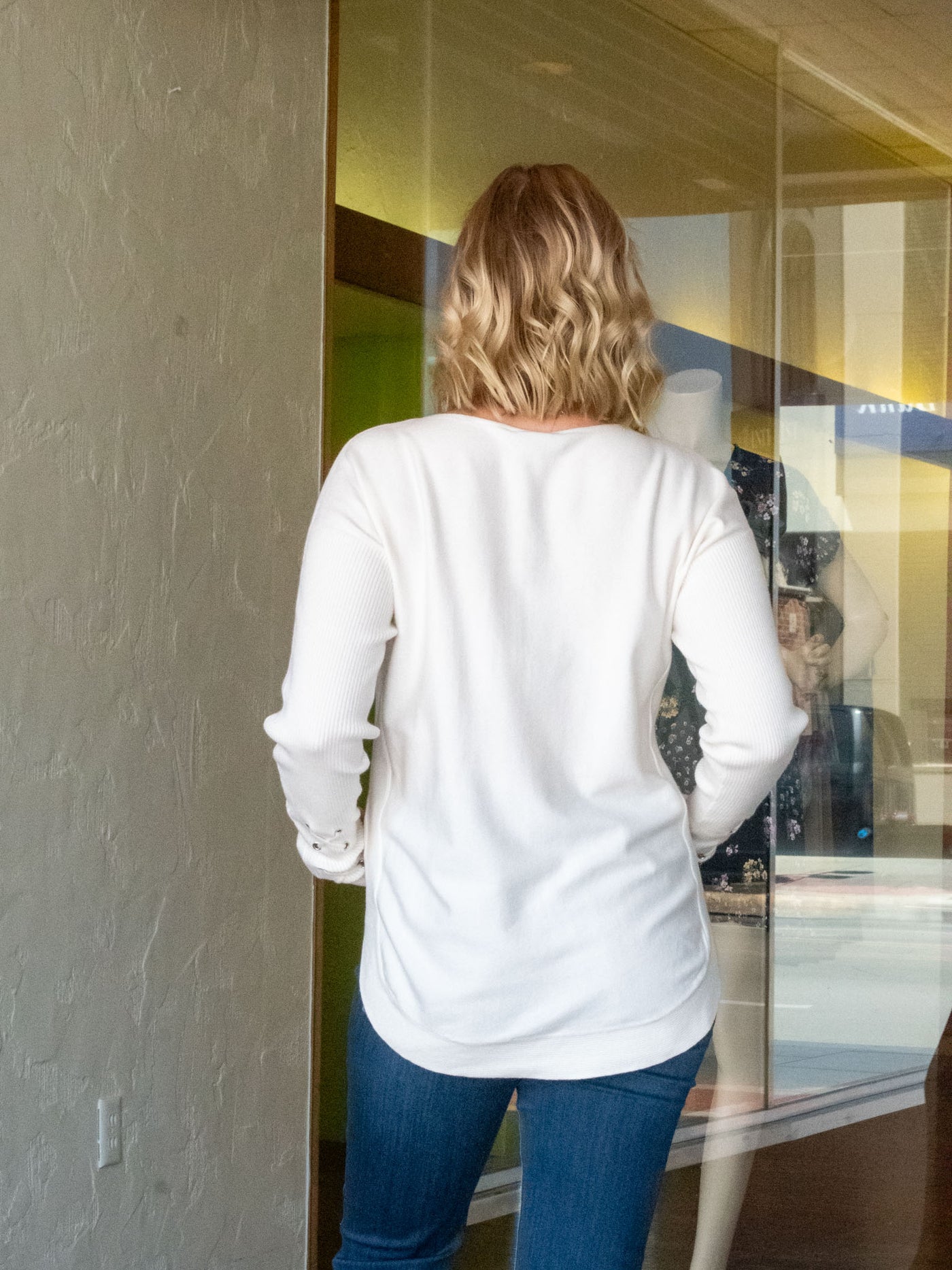 A model wearing a white sweater with a lace-up detail sleeve. The model has it paired with a dark wash flared jean.
