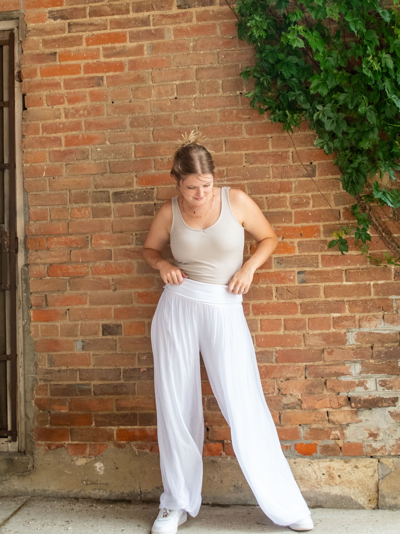 A model wearing a cream ribbed cropped tank top. She has paired it with a pair of flowy silk pants and white sneakers.