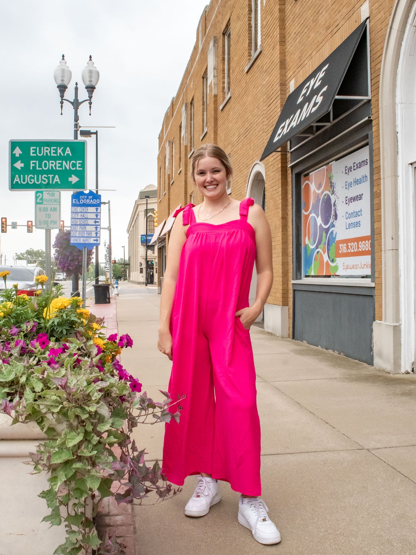 A model wearing a hot pink, wide leg jumpsuit with a sweetheart neckline and bow sleeve details. The model has paired it with white sneakers.