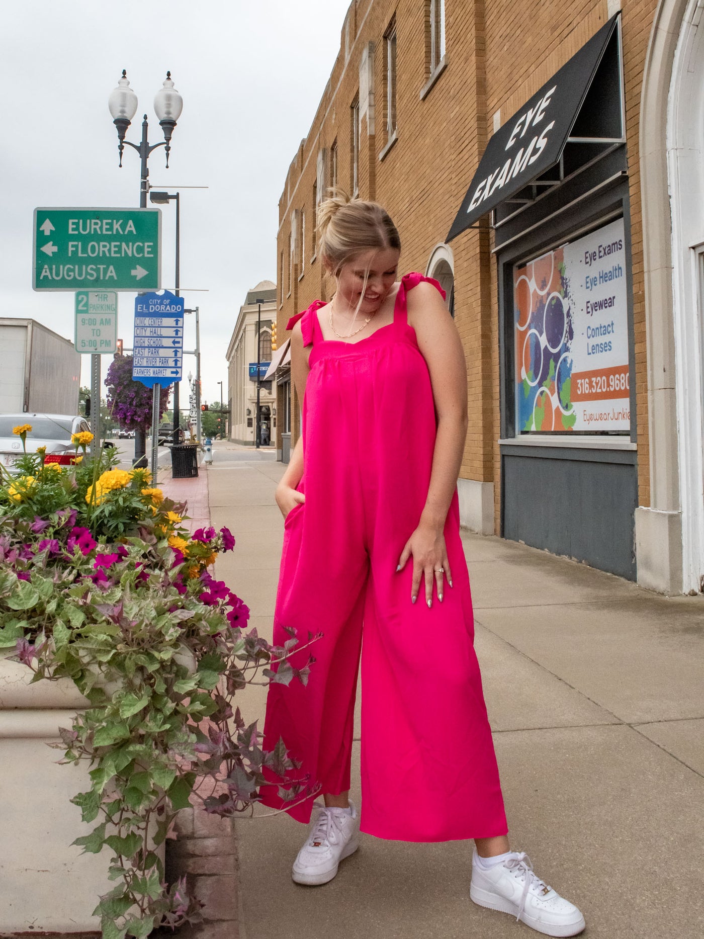 A model wearing a hot pink, wide leg jumpsuit with a sweetheart neckline and bow sleeve details. The model has paired it with white sneakers.