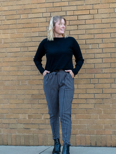 A model wearing a pair of black and white houndstooth joggers. The model has them paired with a black sweater and black booties.
