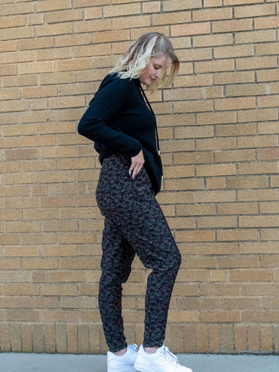 A model wearing a pair of printed joggers with black, grey, and burgundy in them. The model paired it with a black hooded top and white sneakers.