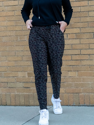 A model wearing a pair of printed joggers with black, grey, and burgundy in them. The model paired it with a black hooded top and white sneakers.