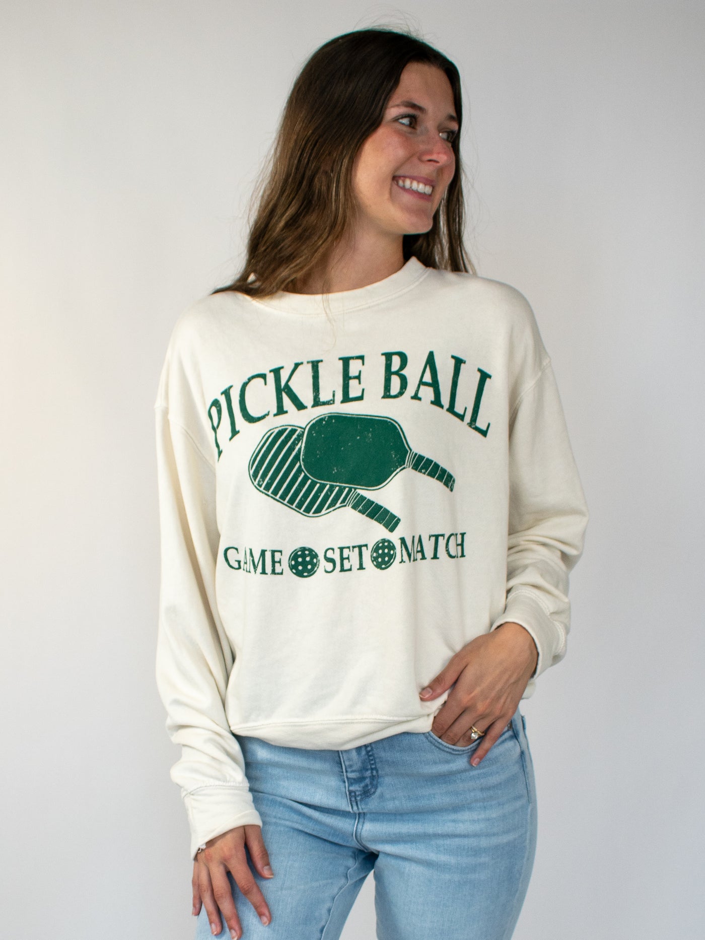 Model wearing a cream crewneck sweatshirt with green lettering that says Pickle Ball and Game, Set, Match with a pickle ball paddle graphic. 