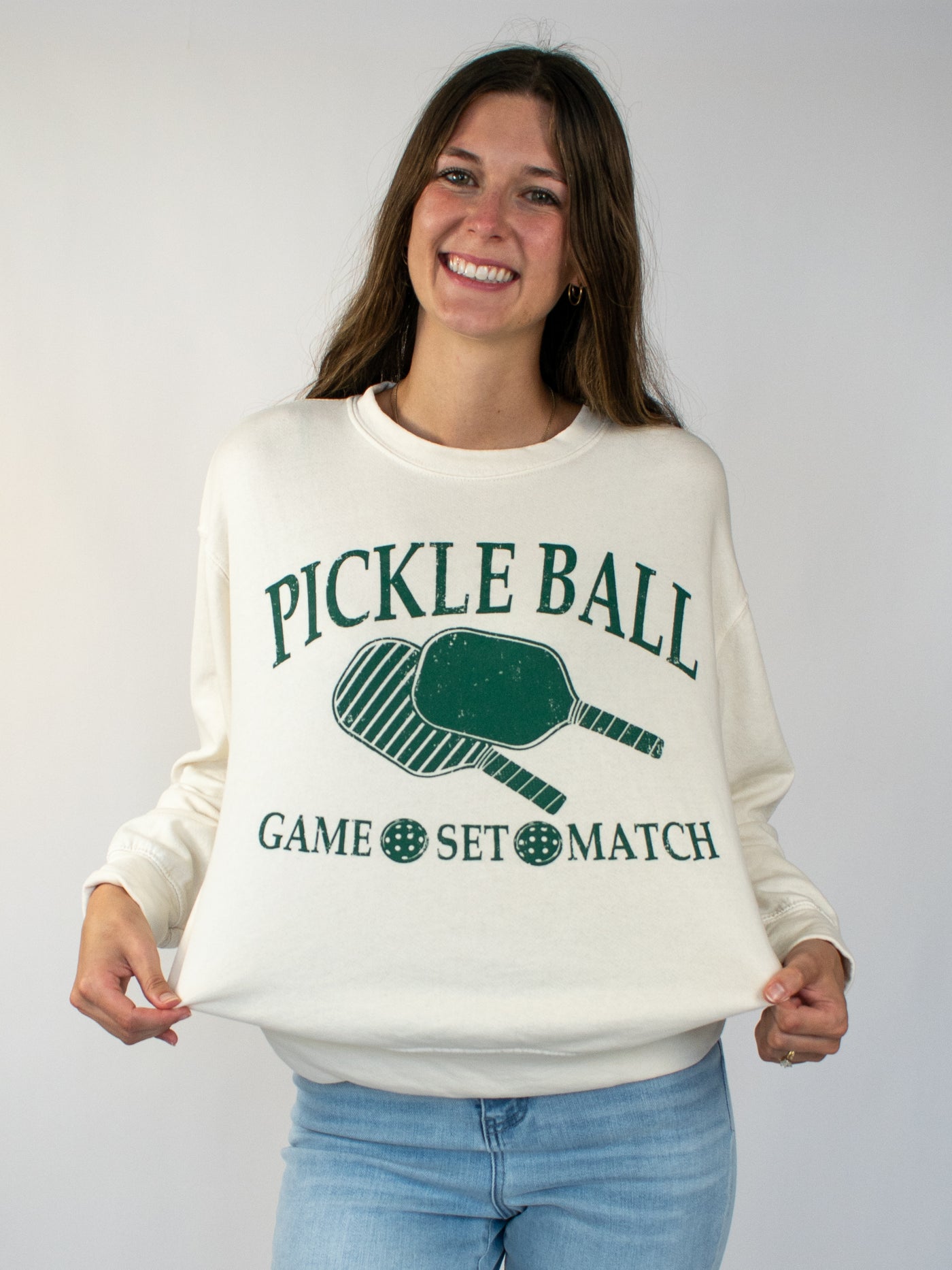 Model wearing a cream crewneck sweatshirt with green lettering that says Pickle Ball and Game, Set, Match with a pickle ball paddle graphic. 