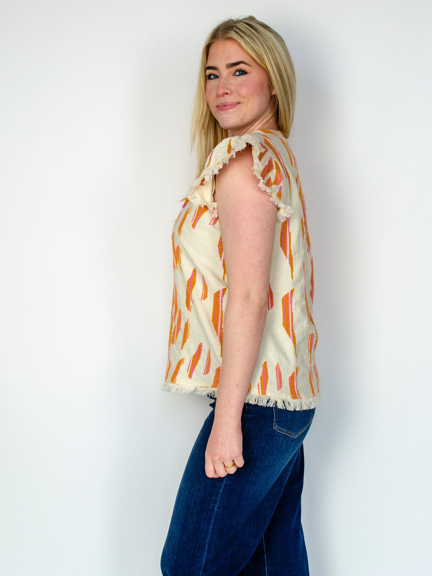 Model is wearing a feminine blouse with frayed hemming on the sleeves and abstract apricot colored detail with a white base color. The blouse is paired with blue jeans. 
