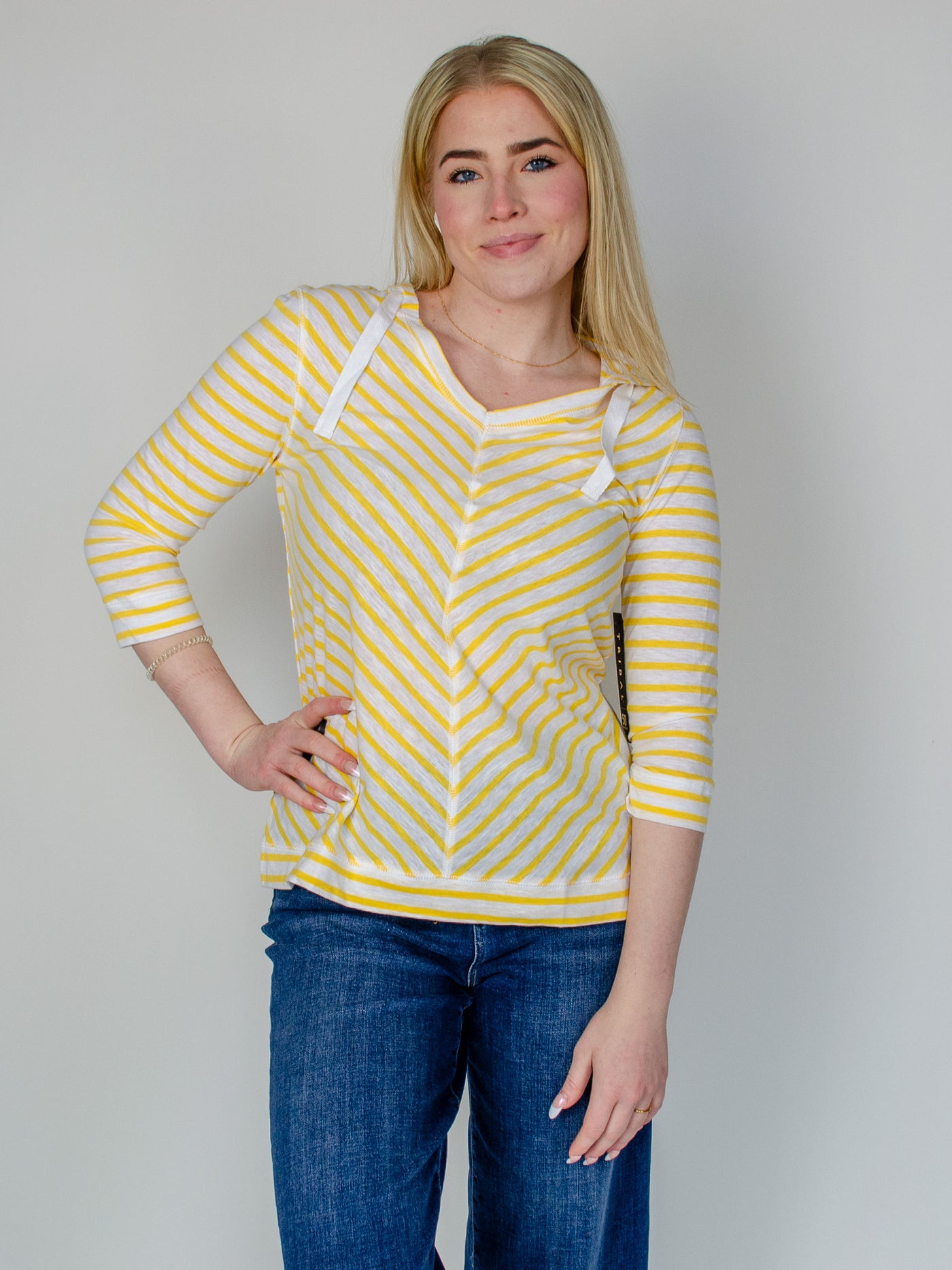 Model is wearing a striped yellow and white quarter sleeve hooded pullover with white drawstring details. Top is paired with dark wash blue jeans. 
