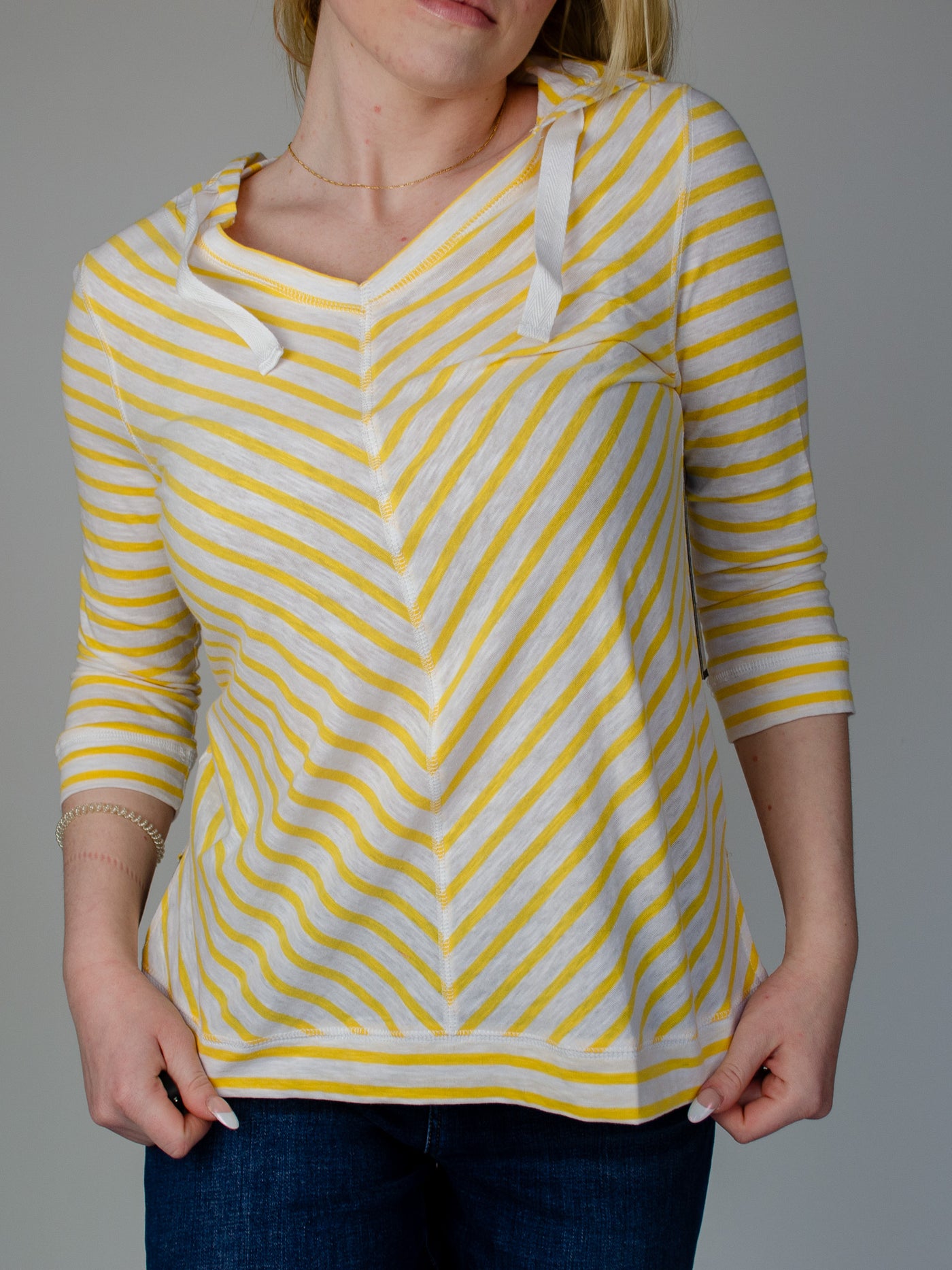 Model is wearing a striped yellow and white quarter sleeve hooded pullover with white drawstring details. Top is paired with dark wash blue jeans.