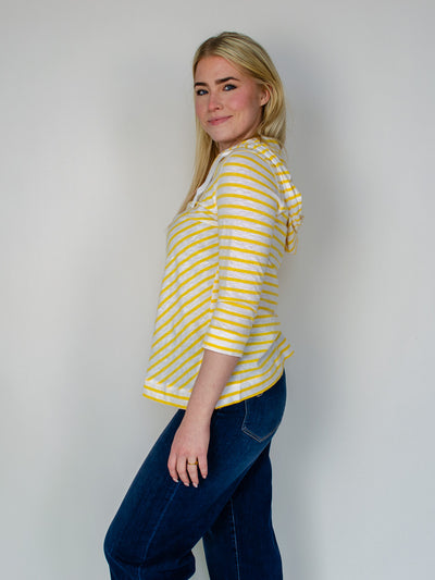 Model is wearing a striped yellow and white quarter sleeve hooded pullover with white drawstring details. Top is paired with dark wash blue jeans.