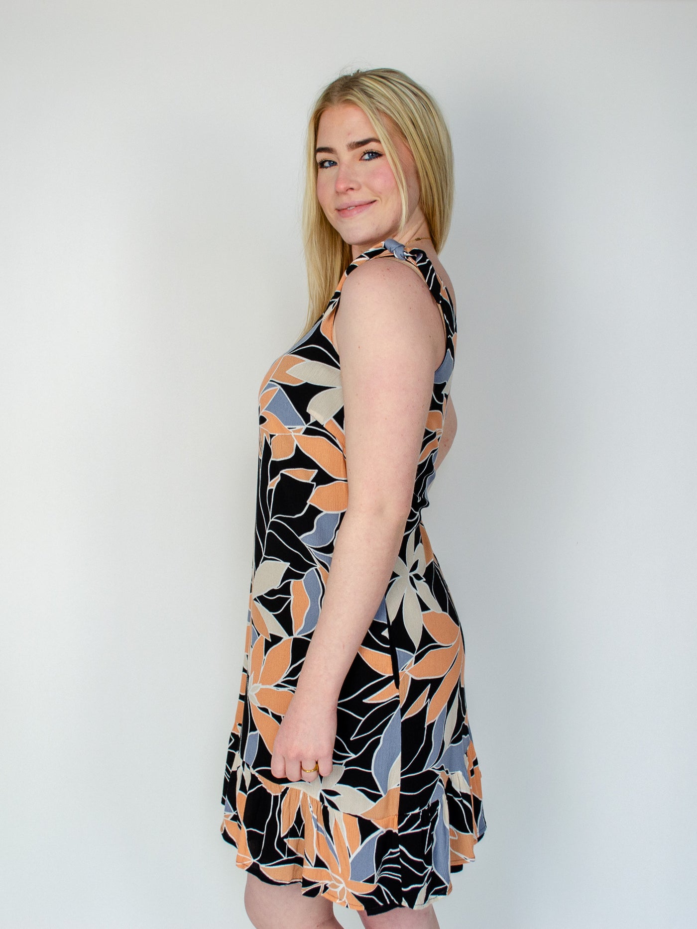Model is wearing a midi dress with a orange, black, and blue colored floral print. The dress has a tie detail on the straps and is reversible and the other side is all black. Dress is paired with white sneakers. 