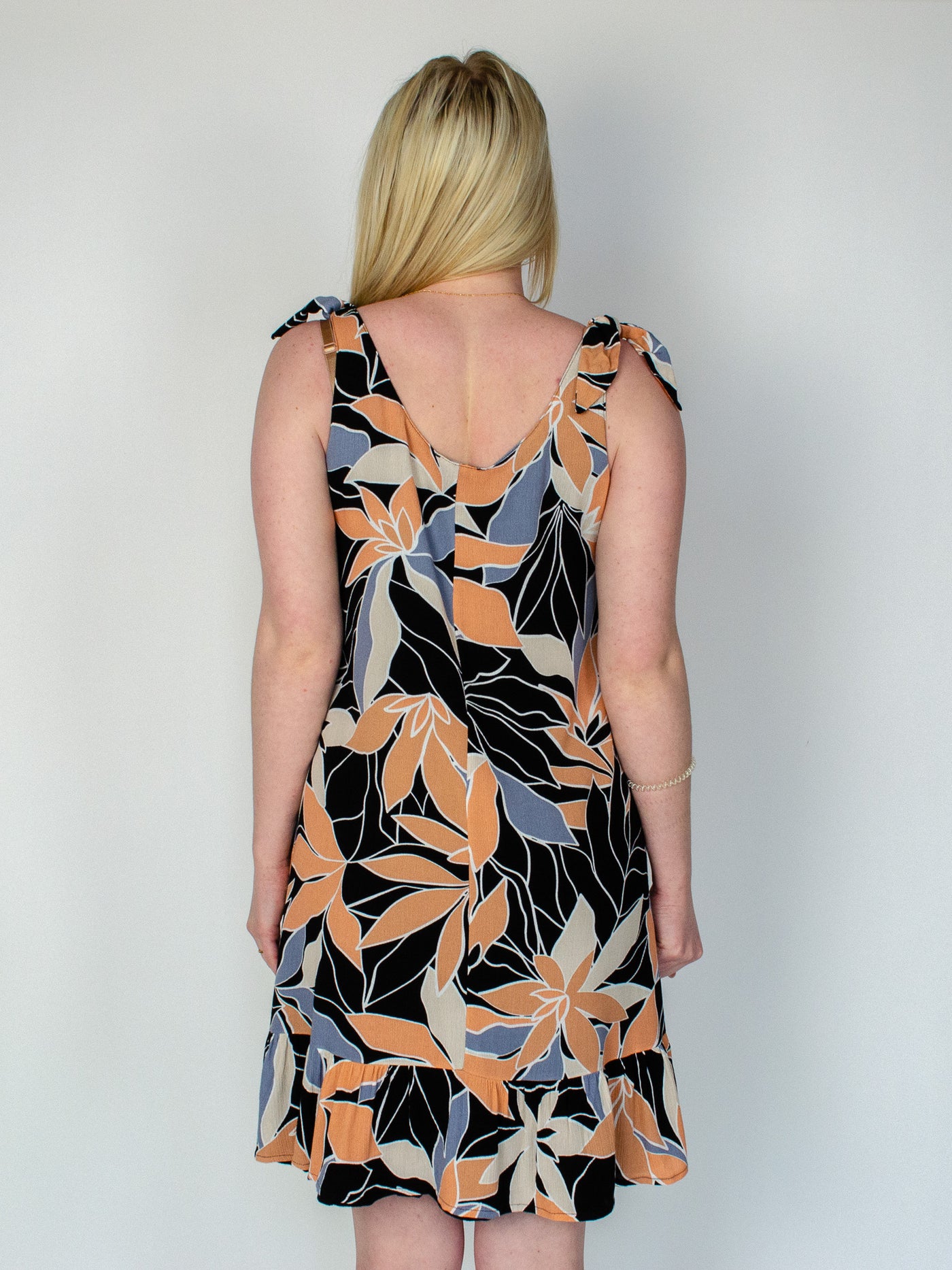 Model is wearing a midi dress with a orange, black, and blue colored floral print. The dress has a tie detail on the straps and is reversible and the other side is all black. Dress is paired with white sneakers. 