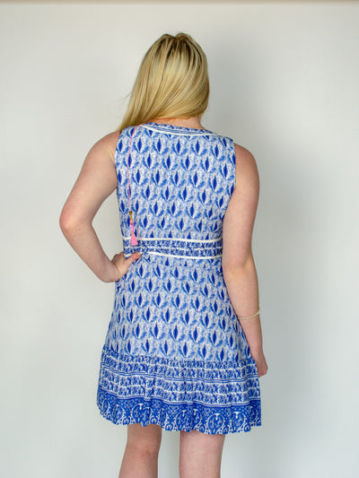 Model is wearing a white and blue patterned midi dress with a V neck neckline and pink tassel details. Dress is paired with white sneakers.  