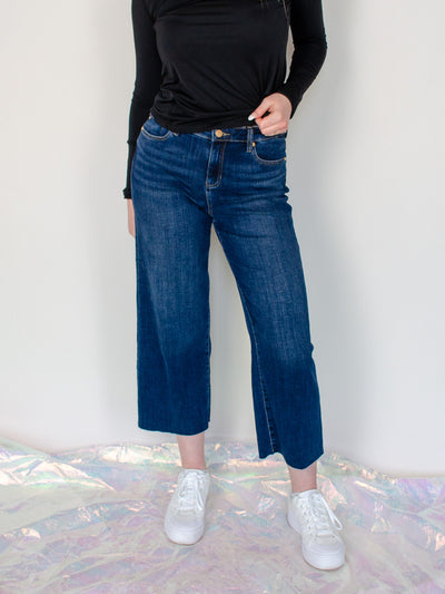 Modeling is wearing a dark wash blue jean that is 3/4th leg length. Jeans are paired with a black fitted longsleeve.