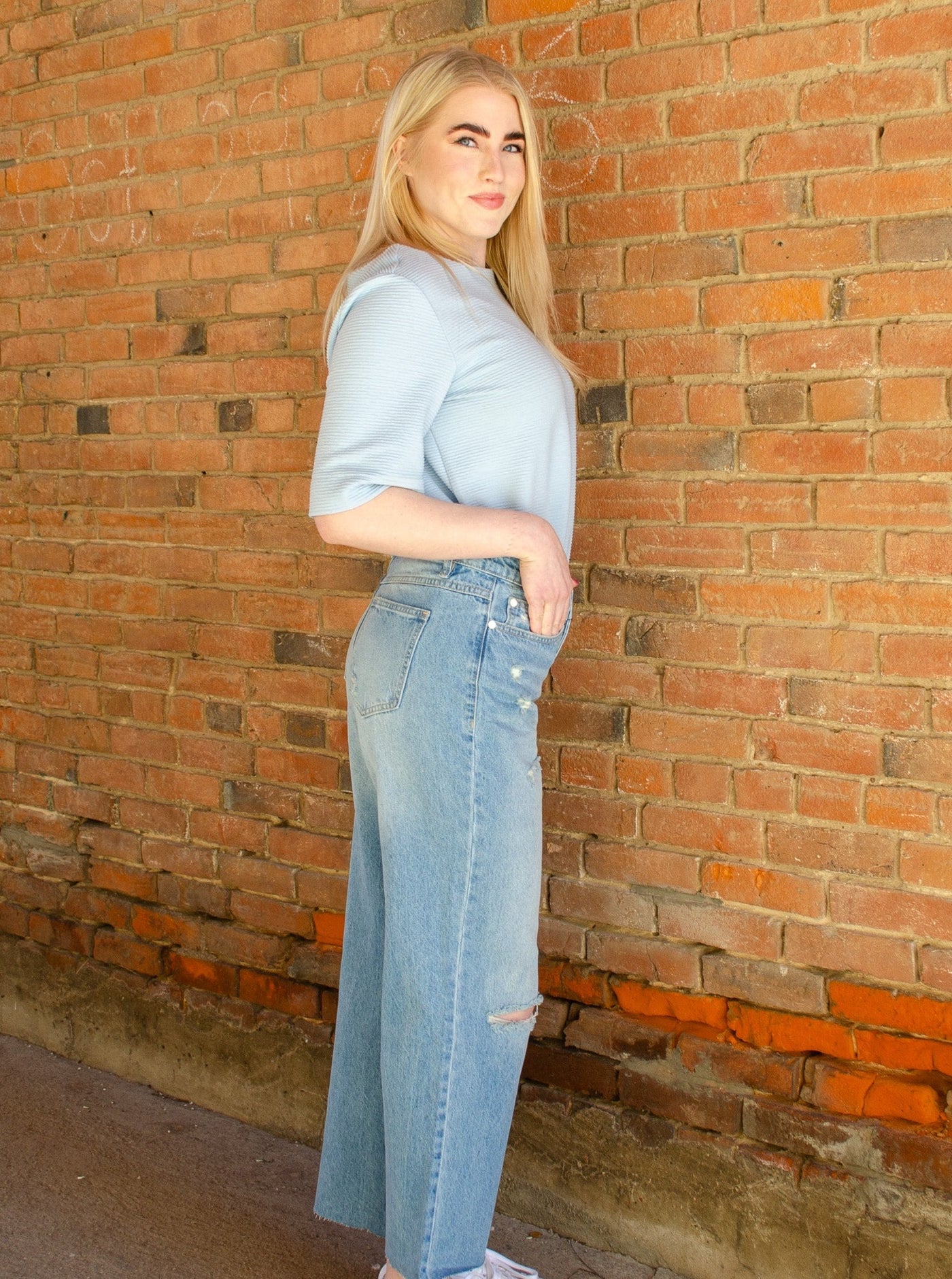 Model is wearing a distressed wide leg boyfriend jean. Jeans are paired with a blue blouse and sneakers. 