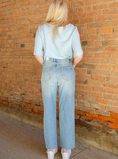 Model is wearing a distressed wide leg boyfriend jean. Jeans are paired with a blue blouse and sneakers. 