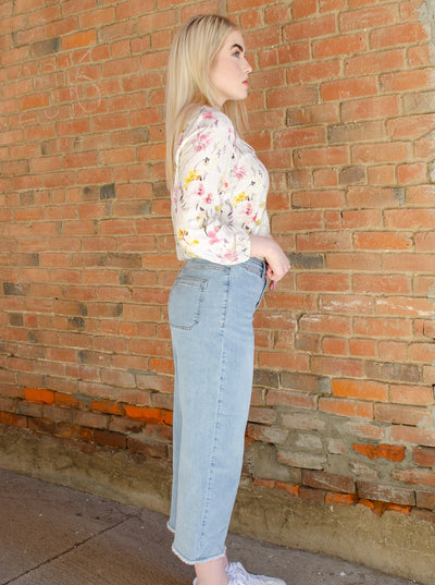 Model is wearing a high waisted light wash button up denim jean with frayed hemming at the ankles. Jeans are worn with a floral blouse.
