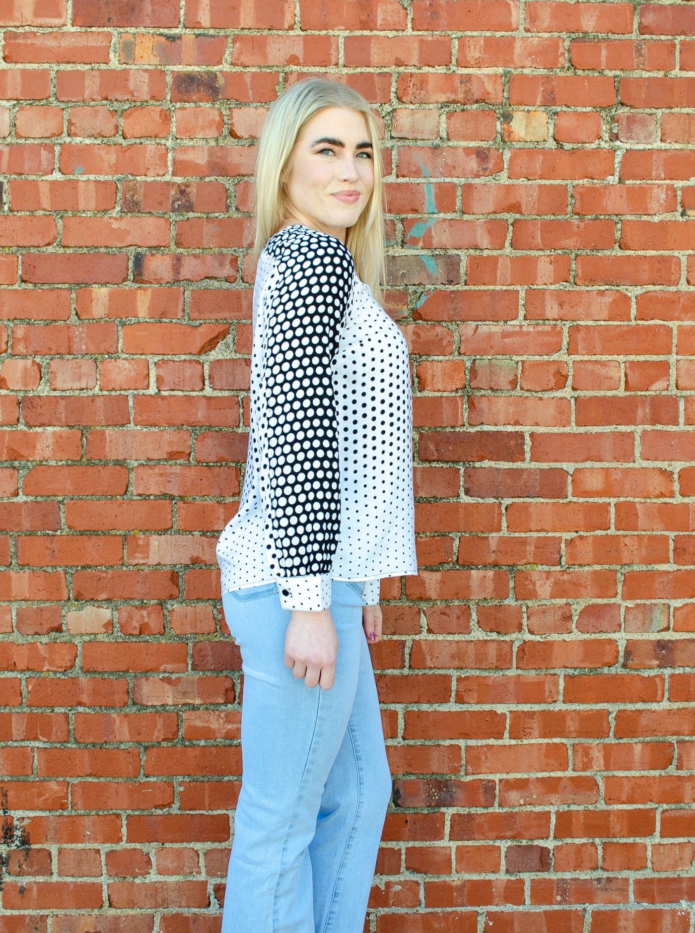 Model is wearing a long sleeved black and white polka dot blouse with puffed sleeves and a v-neck detail on the back of the neckline. 