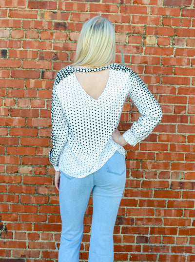 Model is wearing a long sleeved black and white polka dot blouse with puffed sleeves and a v-neck detail on the back of the neckline. 